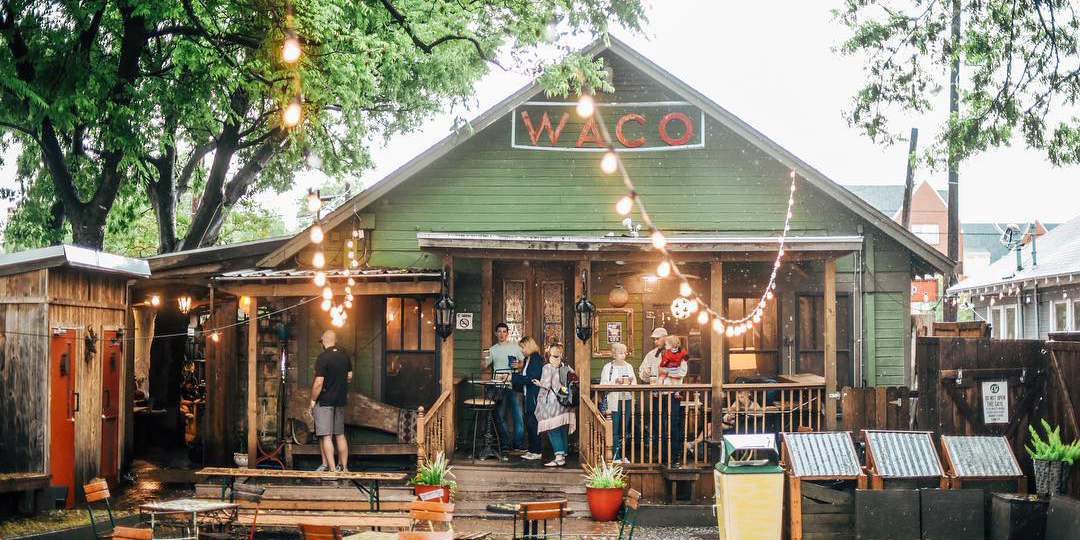 Image of Common Grounds, Waco. The building is green with a wooden porch where several people are gathered. It is surrounded by trees and has hanging lights. The backyard has several picnic tables, and a sign saying Waco is red metal letters hangs from the eaves. 