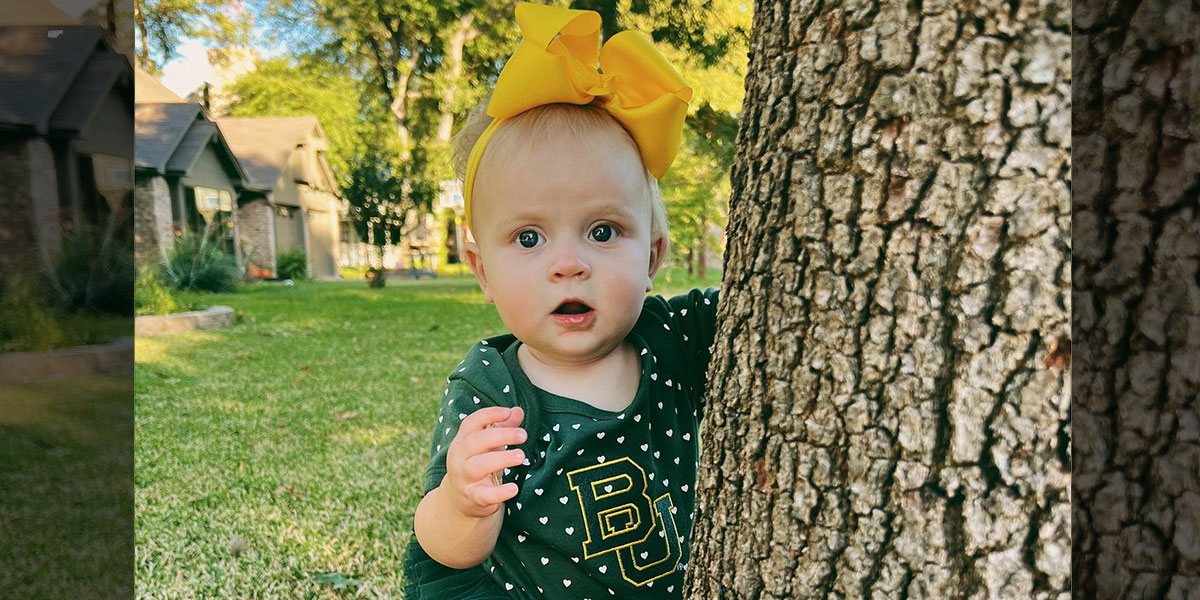 A little girl in a Baylor onesie and yellow bow peers out from around a tree