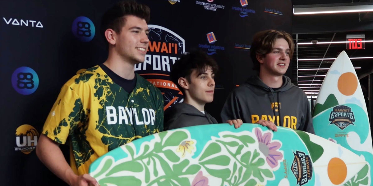 Three Baylor Esports team members stand together in front of a Hawai‘i Esports Invitational backdrop, holding two large surfboard-shaped trophies featuring floral artwork created by a Native Hawaiian artist. Two of the team members wear Baylor apparel, and the group poses to celebrate winning the event.